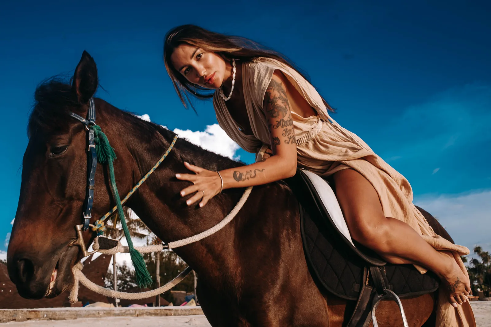 Woman in flowing beige outfit leaning on a brown horse on a sunlit beach