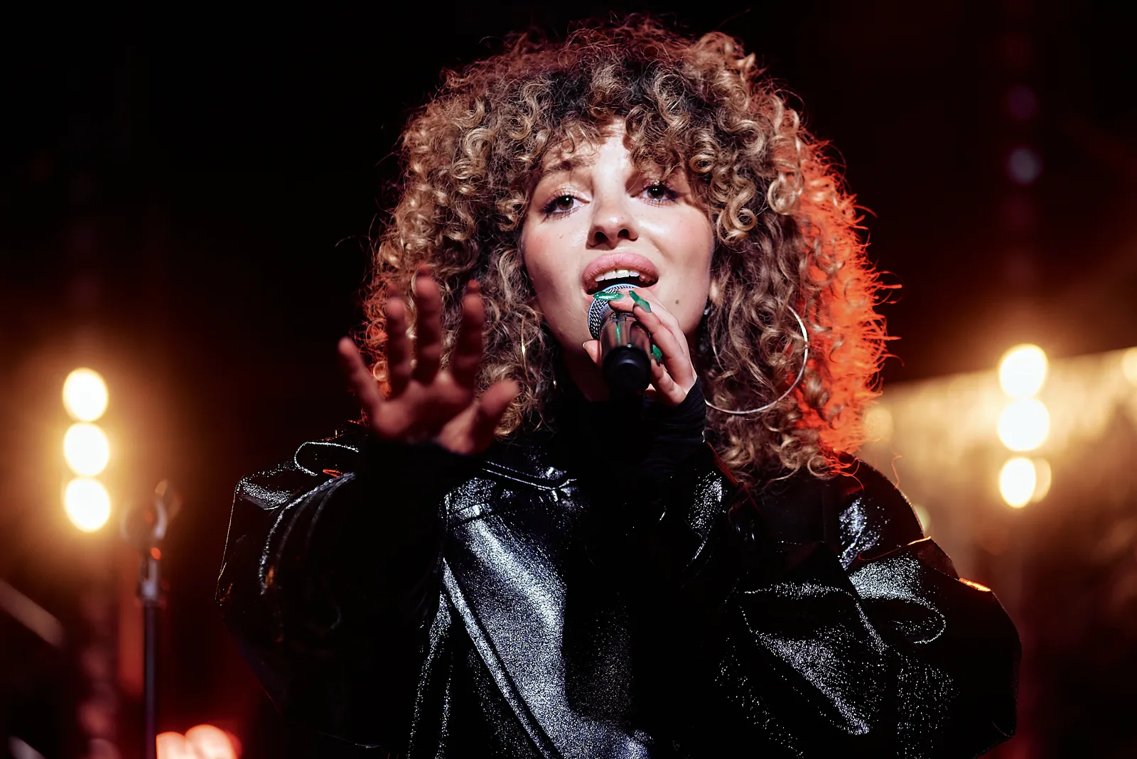 Curly-haired singer in a leather jacket performing under warm stage light