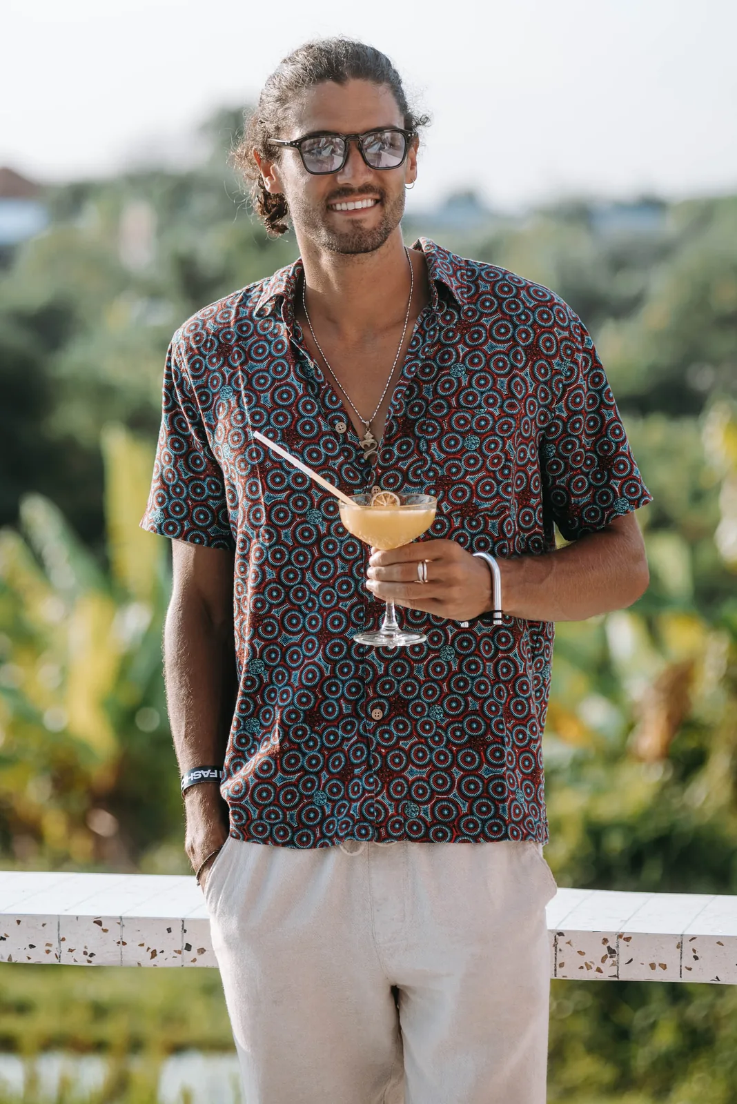 Smiling man in a patterned shirt holding a cocktail on a terrace